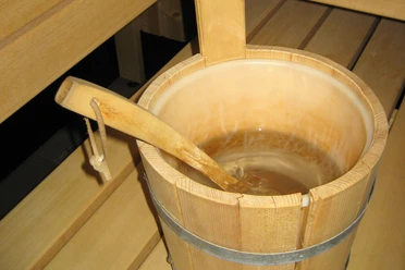 Sauna (3)bearb.jpg Ein hölzerner Saunakübel mit Wasser steht auf einer Holzbank neben einer Saunaaufguss-Kelle.
