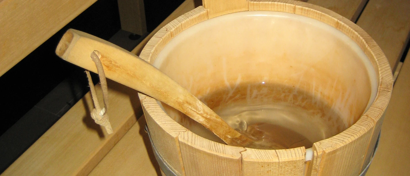 Sauna (3)bearb.jpg Ein hölzerner Saunakübel mit Wasser steht auf einer Holzbank neben einer Saunaaufguss-Kelle.