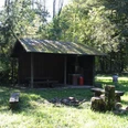 Grillplatz Frangenberg Holzhütte im Wald mit Moos bedecktem Dach, umgeben von grüner Wiese und schattenspendenden Bäumen.