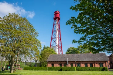 Campener Leuchtturm Rot-weißer Campener Leuchtturm hinter Backsteingebäude, umgeben von grünen Bäumen und blauem Himmel