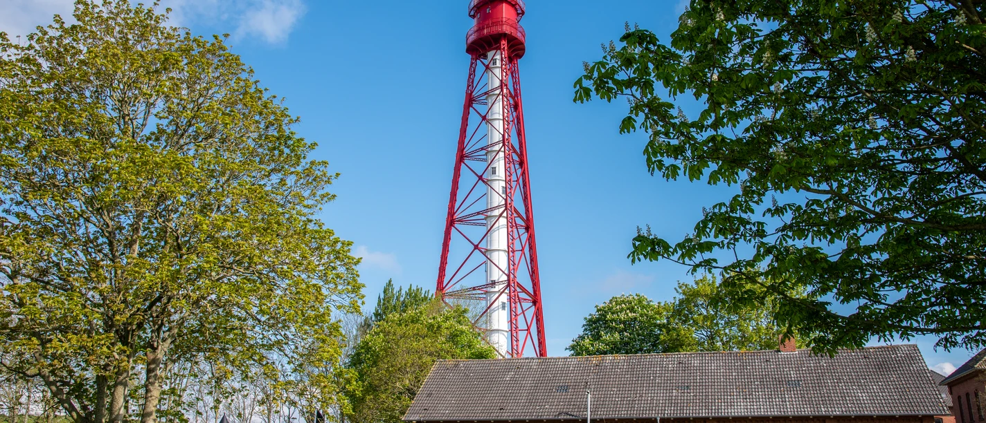 Campener Leuchtturm Rot-weißer Campener Leuchtturm hinter Backsteingebäude, umgeben von grünen Bäumen und blauem Himmel