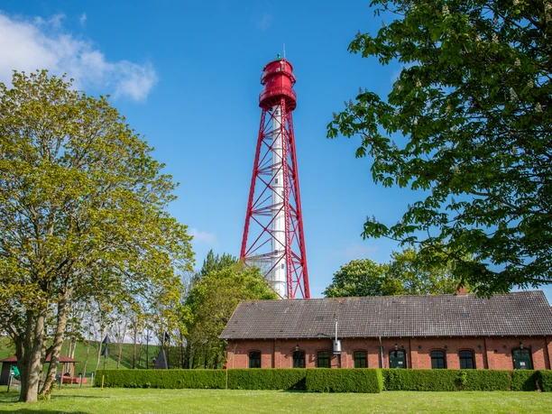 Campener Leuchtturm Rot-weißer Campener Leuchtturm hinter Backsteingebäude, umgeben von grünen Bäumen und blauem Himmel