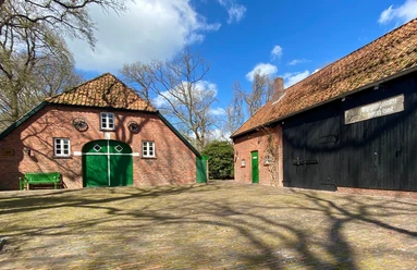 Heinrich-Kunst-Haus außen.jpg Das Bild zeigt eine grüne Parkbank vor zwei rustikalen Gebäuden. Diese haben Backsteinwände, grüne Türen und Fenster.