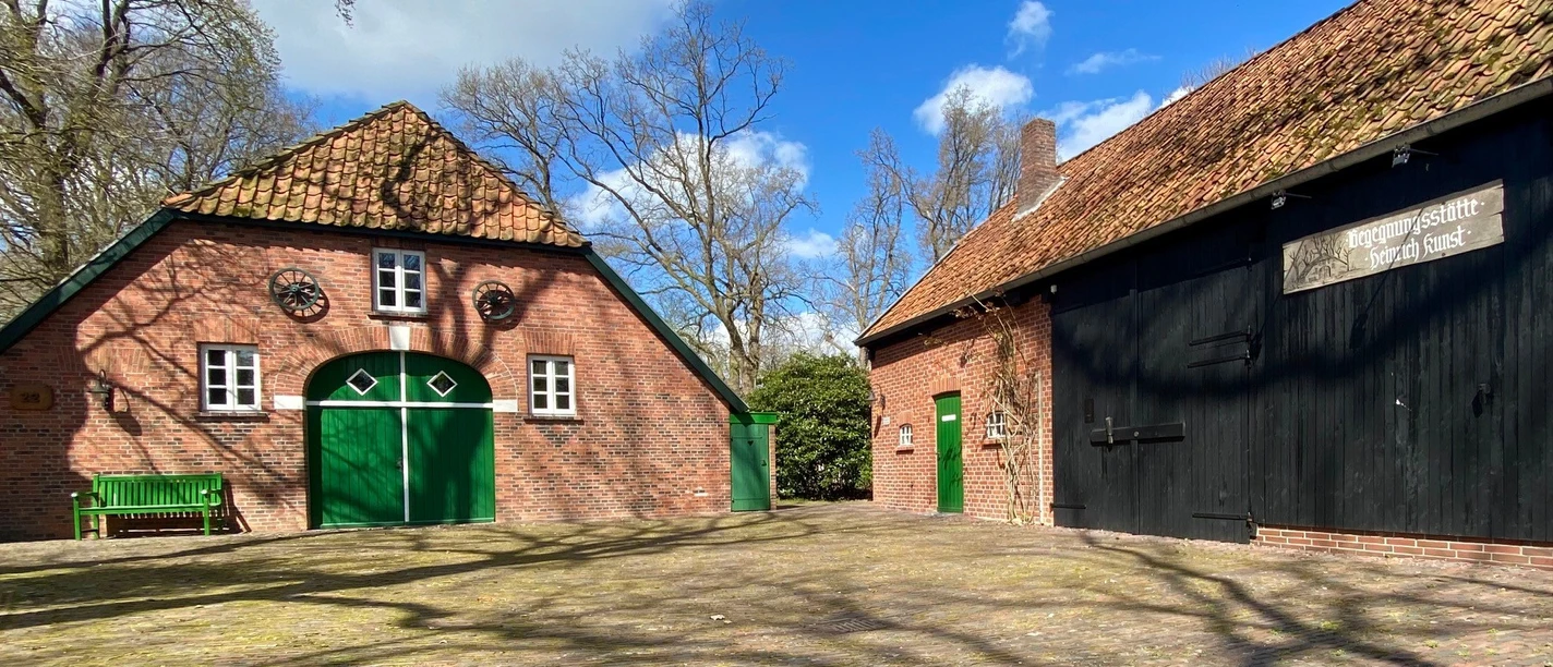 Heinrich-Kunst-Haus außen.jpg Das Bild zeigt eine grüne Parkbank vor zwei rustikalen Gebäuden. Diese haben Backsteinwände, grüne Türen und Fenster.