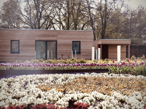 Ein modernes, hölzernes Gebäude steht hinter farbenprächtigen Blumenbeeten in voller Blüte.