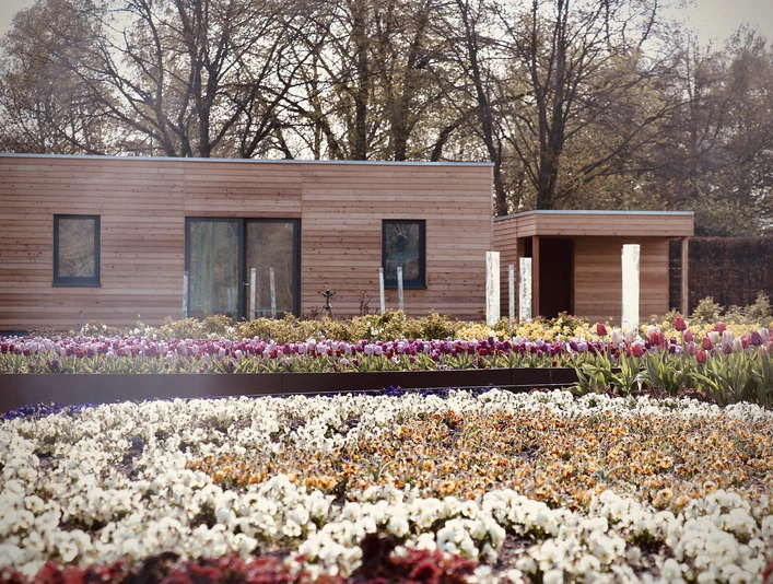 IMG_1366.jpeg Ein modernes, hölzernes Gebäude steht hinter farbenprächtigen Blumenbeeten in voller Blüte.