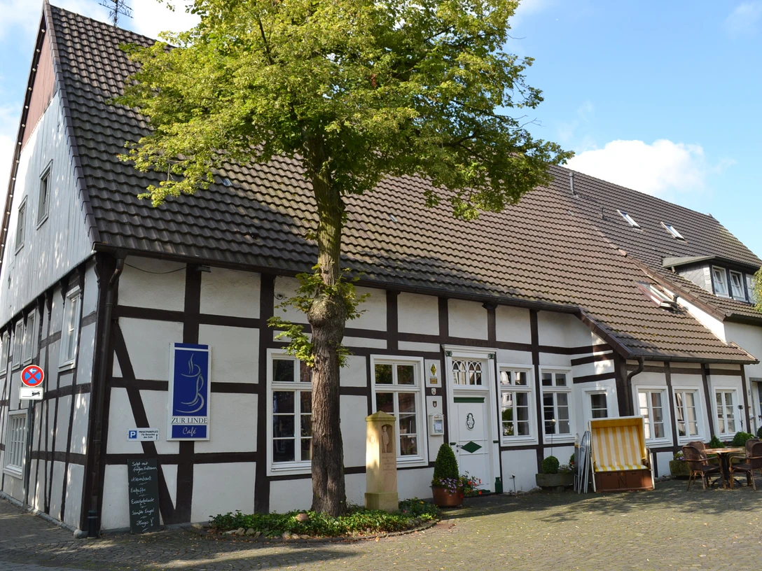 Café zur Linde Langenberg 1.JPG Fachwerkhaus mit Baum im Vordergrund, blauem Schild und gemütlicher Sitzgelegenheit außen.