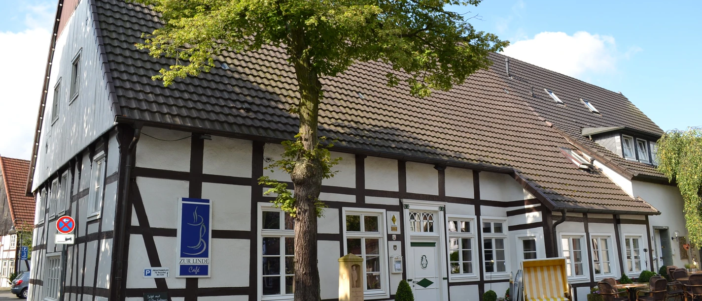 Café zur Linde Langenberg 1.JPG Fachwerkhaus mit Baum im Vordergrund, blauem Schild und gemütlicher Sitzgelegenheit außen.