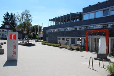Touristinfo Nümbrecht Modernes Gebäude mit Glasfront und orangefarbenem Eingang. Vorplatz mit Bänken und Kreisverkehr.