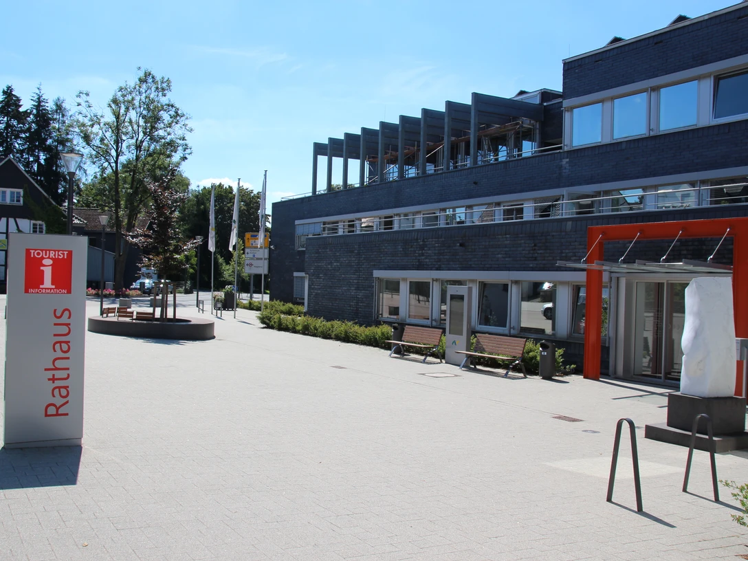 Touristinfo Nümbrecht Modernes Gebäude mit Glasfront und orangefarbenem Eingang. Vorplatz mit Bänken und Kreisverkehr.