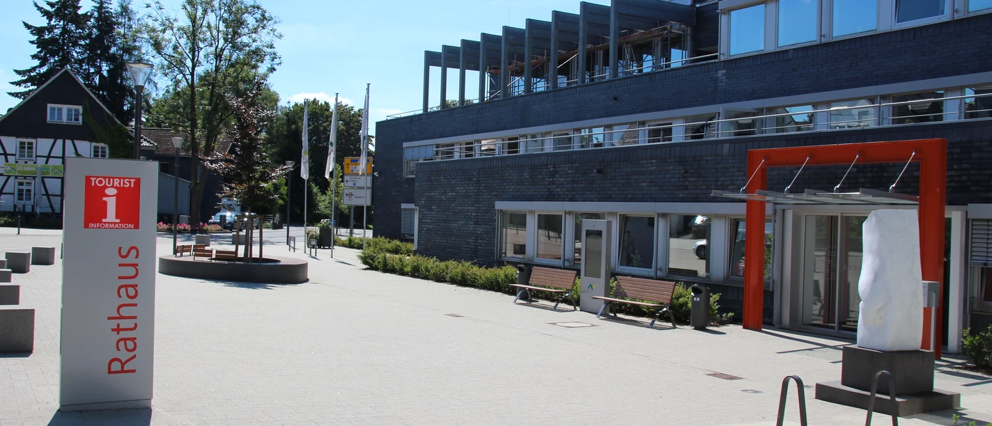 Touristinfo Nümbrecht Modernes Gebäude mit Glasfront und orangefarbenem Eingang. Vorplatz mit Bänken und Kreisverkehr.