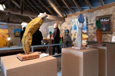 Holzskulpturen stehen auf Sockeln in einem kunstvoll beleuchteten Raum mit Besuchenden im Hintergrund.