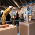 Holzskulpturen stehen auf Sockeln in einem kunstvoll beleuchteten Raum mit Besuchenden im Hintergrund.