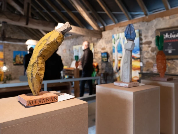 Atelier Schoenen (Foto Stefan Braun)_kl.jpg Holzskulpturen stehen auf Sockeln in einem kunstvoll beleuchteten Raum mit Besuchenden im Hintergrund.