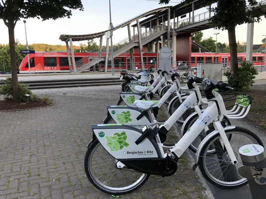 Mobilstation Bergisches e-Bike Bahnhof Overath Fahrräder von "Bergisches e-Bike" an einem Bahnhof, mit rotem Zug im Hintergrund.