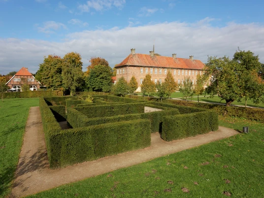 Historisches Herrenhaus mit rotem Ziegeldach und Rosengärtchen im Vordergrund, umgeben von Bäumen.