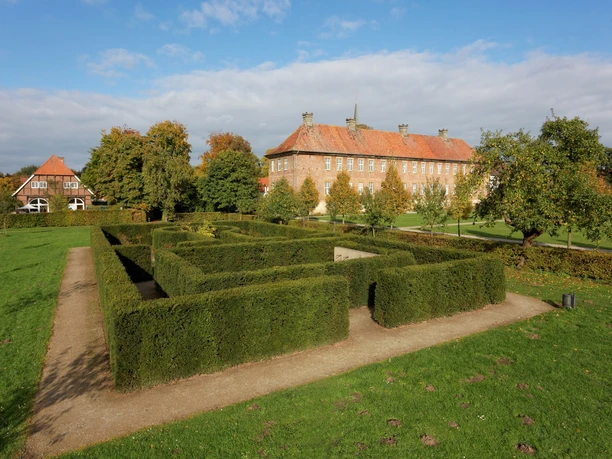 Historisches Herrenhaus mit rotem Ziegeldach und Rosengärtchen im Vordergrund, umgeben von Bäumen.