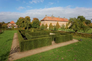 Labyrinthgarten Kloster Clarholz Historisches Herrenhaus mit rotem Ziegeldach und Rosengärtchen im Vordergrund, umgeben von Bäumen.