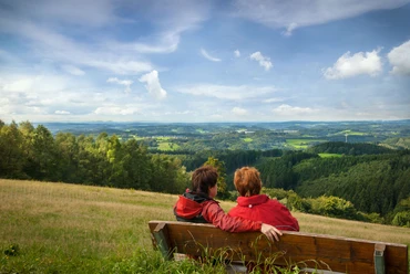 Reichshof-Blockhaus Zwei Personen in roten Jacken sitzen auf einer Bank und blicken in eine weite, grüne Landschaft.