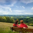 Reichshof-Blockhaus Zwei Personen in roten Jacken sitzen auf einer Bank und blicken in eine weite, grüne Landschaft.
