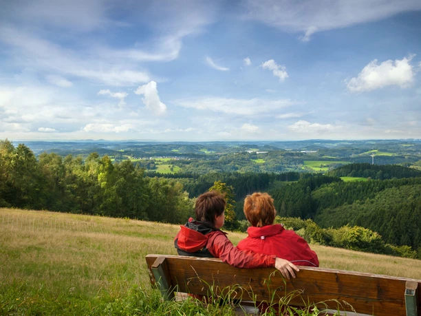 Reichshof-Blockhaus Zwei Personen in roten Jacken sitzen auf einer Bank und blicken in eine weite, grüne Landschaft.
