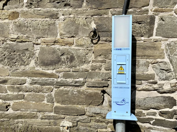 E-Bike Ladesäule Mühle Nespen E-Ladestation an einer alten Steinmauer befestigt, mit zwei Steckdosen und einem Warnsymbol versehen.
