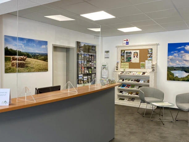 Kur- und Touristinfo Reichshof Empfangsbereich mit Informationsbroschüren, modernen Möbeln, Landschaftsfotos und einem Begrüßungsschild.