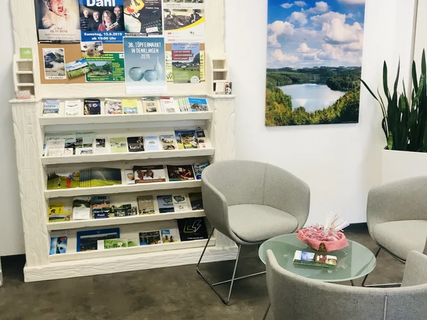 Kur- und Touristinfo Reichshof Warteraum mit grauen Sesseln, Zeitschriftenwand und einem Landschaftsfoto an der weißen Wand.