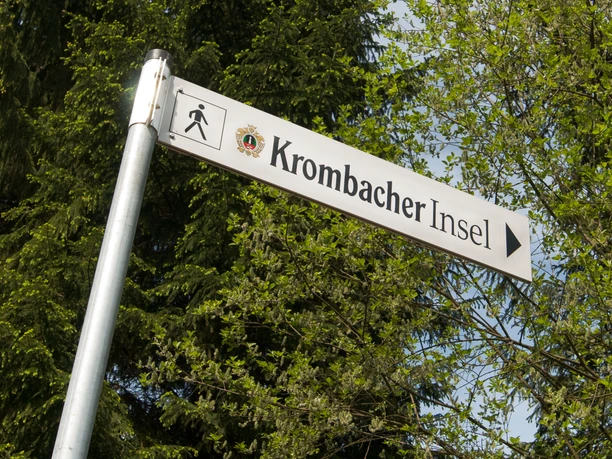 Krombacher-Insel Wegweiser zur Krombacher Insel, umgeben von grünen Bäumen im Hintergrund, zeigt Fußgänger-Symbol.