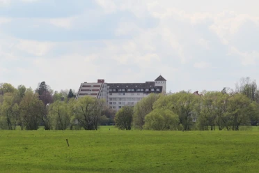 Eine Kurklinik hinter grünen Bäumen, im Hintergrund ein leicht bewölkter, blauer Himmel.