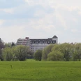 Kurklinik Bad Hopfenberg Eine Kurklinik hinter grünen Bäumen, im Hintergrund ein leicht bewölkter, blauer Himmel.