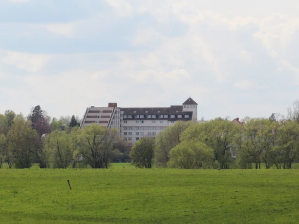 Kurklinik Bad Hopfenberg Eine Kurklinik hinter grünen Bäumen, im Hintergrund ein leicht bewölkter, blauer Himmel.
