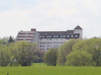 Kurklinik Bad Hopfenberg Eine mehrstöckige Klinik mit schlankem Turm erhebt sich hinter einer grünen Baumreihe im Frühling.