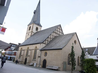 Eine historische Kirche mit hohem Turm und Fachwerk, umgeben von urbaner Architektur und Spaziergängern.