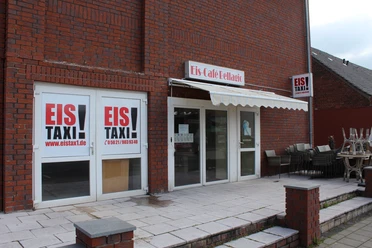Eistaxi Eiscafé mit roter Backsteinfassade, großen Fenstern und einer Terrasse mit gestapelten Stühlen.