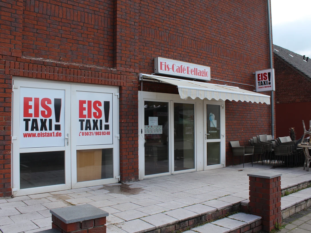 Eistaxi Eiscafé mit roter Backsteinfassade, großen Fenstern und einer Terrasse mit gestapelten Stühlen.