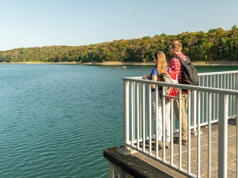 Wahnbachtalsperre Paar genießt Aussicht auf einen ruhigen See mit bewaldeten Ufern bei strahlendem Sonnenschein.