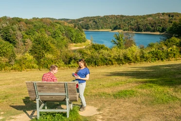 Wahnbachtalsperre Zwei Personen auf einer Bank in einer grünen Landschaft mit einem See im Hintergrund.