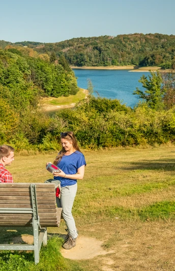 Wahnbachtalsperre Zwei Personen auf einer Bank in einer grünen Landschaft mit einem See im Hintergrund.