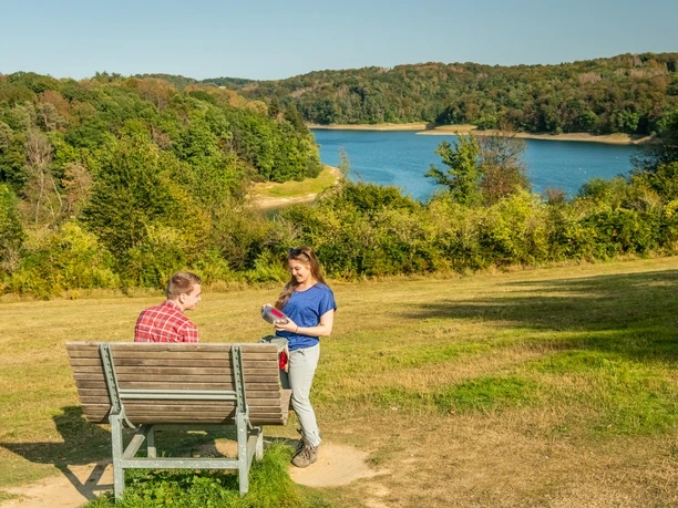 Wahnbachtalsperre Zwei Personen auf einer Bank in einer grünen Landschaft mit einem See im Hintergrund.