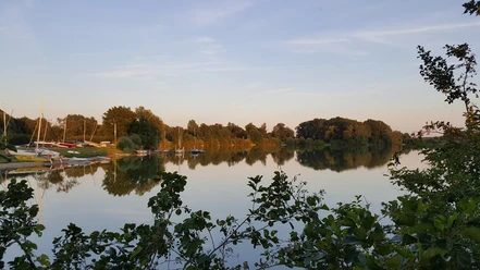 Wassersport am Linteler See Ein ruhiger See mit Segelbooten am Ufer, umgeben von Bäumen, reflektiert den Abendhimmel.
