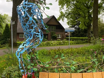 Eichenkönig mit Bank Eine filigrane Metallskulptur mit organischen Strukturen sitzt auf einer Holzbank, umgeben von blühender Vegetation.