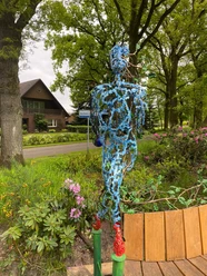 Lieblingsort Klauhörn - Eichenkönig Eine filigrane Skulptur aus Stahl steigt vor dichten, grünen Bäumen empor, umgeben von bunter Bepflanzung. Im Hintergrund erkennt man ein Haus hinter der belebten Natur.