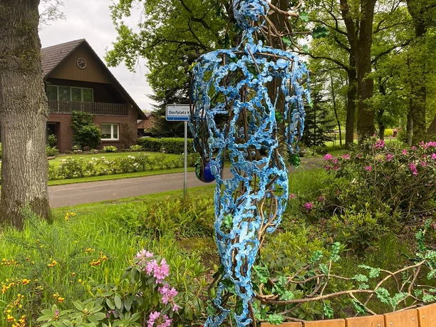 Lieblingsort Klauhörn - Eichenkönig Eine filigrane Skulptur aus Stahl steigt vor dichten, grünen Bäumen empor, umgeben von bunter Bepflanzung. Im Hintergrund erkennt man ein Haus hinter der belebten Natur.