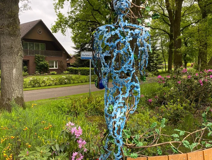 Lieblingsort Klauhörn - Eichenkönig Eine filigrane Skulptur aus Stahl steigt vor dichten, grünen Bäumen empor, umgeben von bunter Bepflanzung. Im Hintergrund erkennt man ein Haus hinter der belebten Natur.