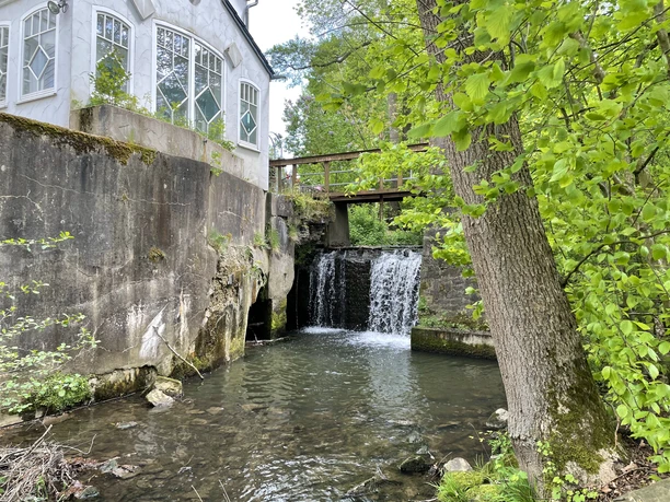 Oud gebouw met waterval en brug, omringd door groene bomen en een stromend beekje.