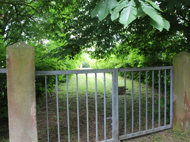 Jüdischer Friedhof Lavelsloh Metallzaun mit Tor, umgeben von üppigem grünem Blattwerk und Baumschatten, führt zu einer Rasenfläche.