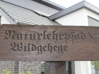 Holzschild "Naturlehrpfad Wildgehege" vor modernem Gebäude mit Solarpanel auf dem Dach.