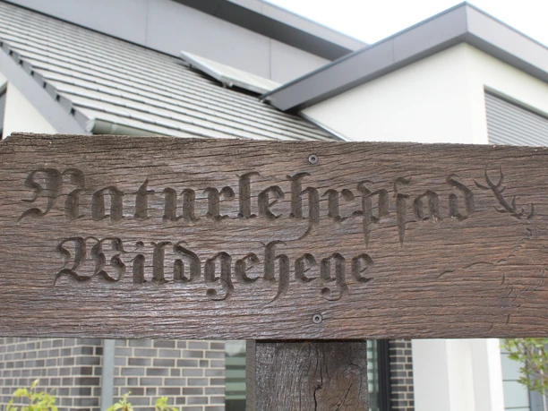 Naturlehrpfad Holzschild "Naturlehrpfad Wildgehege" vor modernem Gebäude mit Solarpanel auf dem Dach.