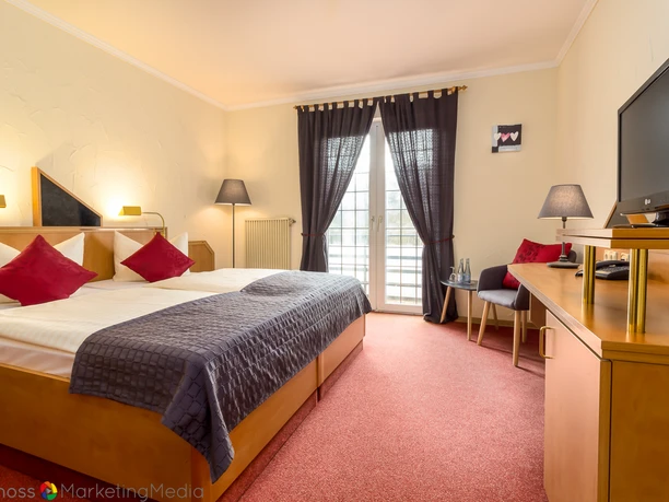 Zimmer (1).jpg Ein helles Hotelzimmer mit Doppelbett, dunklen Akzenten, Schreibtisch und Balkon mit Vorhängen.